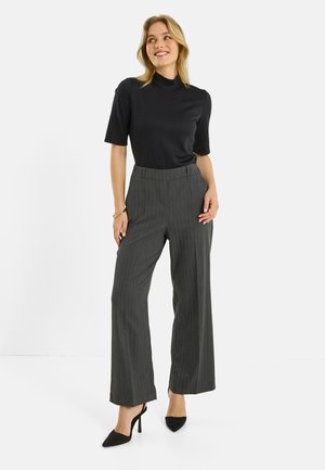 Mujer con una blusa negra de manga corta y cuello alto, pantalones anchos a rayas grises y zapatos negros de tacón alto, de pie sonriendo.