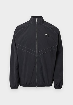 Chaqueta cortavientos de nylon negro con cuello alto, cierre frontal de cremallera y puños elásticos, con costuras contrastantes y un pequeño parche con logotipo.