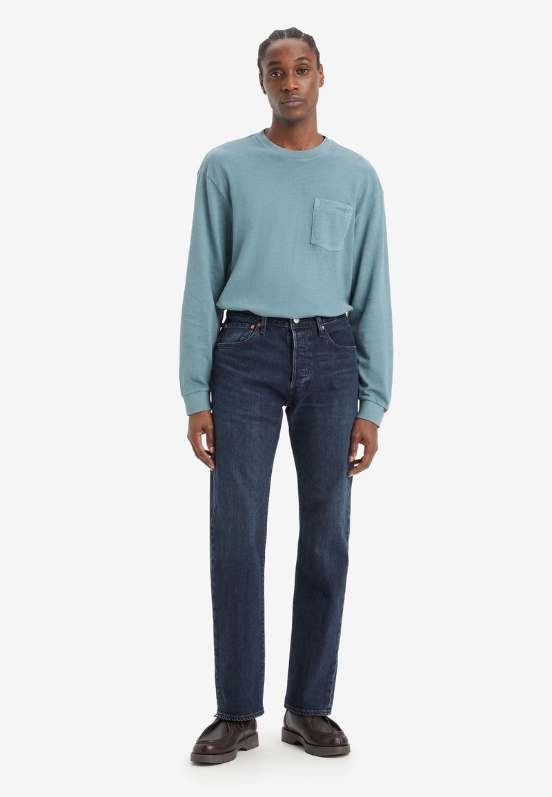 Lange mouwen teal katoenen shirt met een borstzak, gecombineerd met donkerblauwe denim jeans en bruine leren schoenen, staande tegen een effen witte achtergrond.