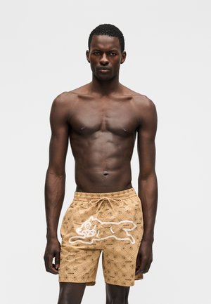 Αθλητικός άνδρας που στέκεται στραμμένος μπροστά, φορώντας μπεζ σορτς με σχέδιο και ένα λευκό καρτουνίστικο ζώο μπροστά, γυμνόφατσα σε απλό φόντο.