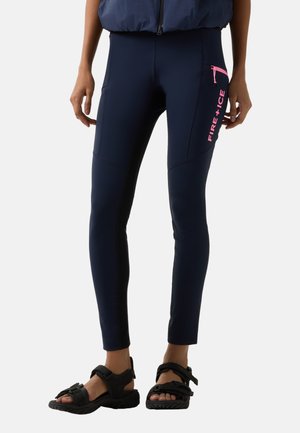 Slim-fit marineblauwe broek met een roze zak met rits en de tekst "FIRE + ICE", gedragen met zwarte sandalen en een marineblauwe jas.