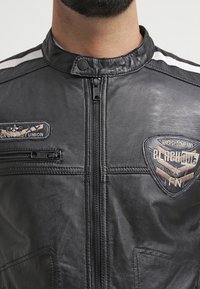 Sort læderjakke med høj krave, lynlås, sidelommer og broderede mærker med skjolddesign og striber.