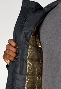 Jachetă exterioară neagră, cu o căptușeală interioară aurie cu model de fagure strălucitor și detalii vizibile ale fermoarului, purtată peste o cămașă gri. O mână apucând jacheta.