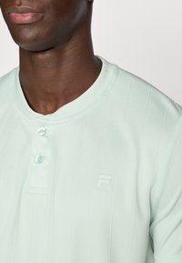 Ανοιχτό πράσινο, υφασμάτινο πόλο με κάθετες ρίγες, δύο κουμπιά και μικρό, ανάγλυφο λογότυπο στο στήθος.