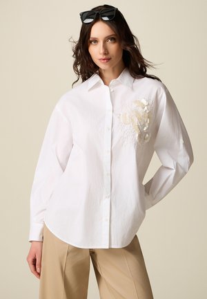 Femme portant une chemise blanche boutonnée avec une broderie florale beige sur la poitrine, un pantalon beige et des lunettes de soleil noires reposant sur sa tête.