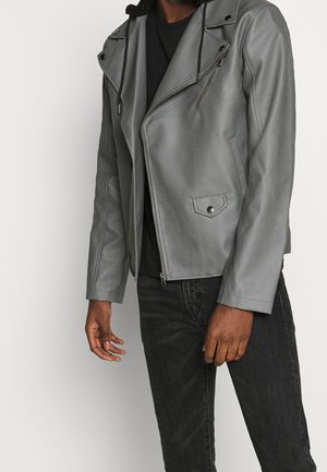 Mężczyzna ubrany w szarą skórzaną kurtkę motocyklową z zamkiem błyskawicznym i kieszenią na zatrzask, w zestawieniu z czarnymi dżinsami i czarną koszulą.