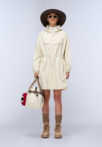 Giacca impermeabile beige chiaro con vita regolabile, maniche lunghe e colletto alto. Il modello tiene in mano una borsa a righe beige e rossa, indossando calzini e stivali marroni.