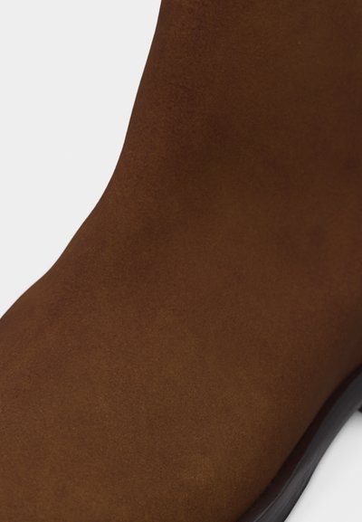 Botte en daim marron à texture lisse avec semelle noire visible sur le bord, sur fond blanc.