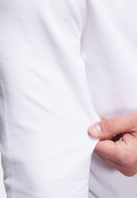Vit texturerad tyg med ett rutmönster, med en hand som greppar ärmen. Materialet verkar lätt och andas.