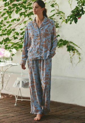 Frau steht barfuß in einem lockeren blauen Schlafanzug mit Blumenmuster neben einem weißen Tisch und grünem Efeu an einer weißen Wand.