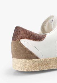 Sneaker bianco con una suola beige testurizzata, un pannello laterale in suede marrone e un accento scintillante in bronzo sul tallone. Parte superiore in pelle liscia.