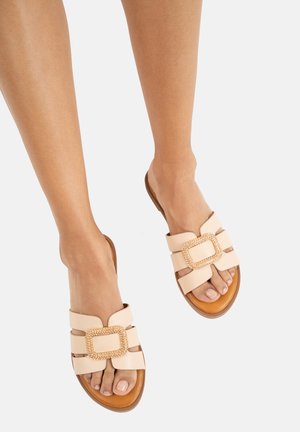Nude slip-on sandals with gold accent buckle, featuring three wide straps and a flat brown sole. Smooth leather material and open toe design.