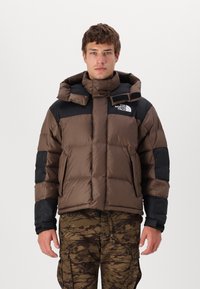 Brun puffjacka med svarta detaljer, med hög krage och huva. Texturerad yta med quiltat mönster. Logotyp på bröstet.