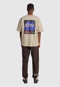 Beige oversized t-shirt med en blå och svart grafisk design tryckt på baksidan, sammansatt med mörkbruna byxor som har vit text och dragkedjefickor.