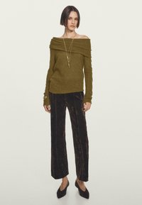 Maglione verde oliva a spalla scoperta in morbido tessuto a maglia, abbinato a pantaloni larghi in velluto scuro e décolleté neri a punta.