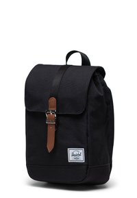 Black fabric backpack with a fold-over flap, brown leather strap and metal buckle, and a white logo patch on the lower front.