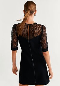 Schwarzes Kleid mit einer Spitzentaille und Ärmeln in Ellenbogenlänge. Der Rücken hat eine Schlüsselöffnungsöffnung und einen Reißverschluss. Glatte Stofftextur.