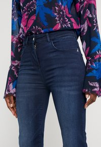 Jeans de mezclilla oscura con corte slim y sutiles cremalleras en los bolsillos, combinados con una blusa de patrón floral colorido con mangas acampanadas.