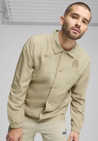 Beige Puma jakke med krave, trykknapper og broderet holdlogo. Glat tekstur og elastiske manchetter.