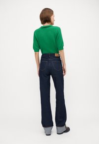 Zelený, rebrovaný pletený polo s krátkymi rukávmi skombinovaný s nohavicami s vysokým pásom v tmavomodrej farbe, ktoré majú hnedú koženú nášivku a prehnuté manžety.