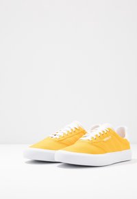 Gula canvas sneakers med vita snören och sulor. Har en texturerad yta, subtila ränder och logotyp på sidan. Låg toppdesign.