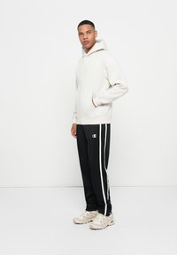 Felpa grigia chiara con tasca a marsupio e coulisse, abbinata a pantaloni della tuta neri con strisce bianche laterali e logo. Scarpe da ginnastica beige.