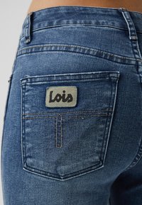 Mørkeblå denimjeans har en baklomme med en grå lapp merket "Lois" med svart skrift og kontrasterende gul søm.