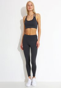 Donkergrijze sportbeha met dunne bandjes en bijpassende high-waisted leggings, gemaakt van soepel, rekbaar materiaal; gecombineerd met witte sportschoenen.