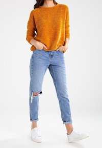 Gestrickter Pullover in senfgelb mit strukturierter Oberfläche, lockere Passform. Hellblaue Jeans mit hoher Taille und einem Riss am Knie, an den Knöcheln aufgerollt. Weiße Turnschuhe.