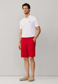 Uomo in piedi con una mano in tasca, che indossa una polo bianca, pantaloncini rossi e scarpe beige senza lacci su sfondo neutro.