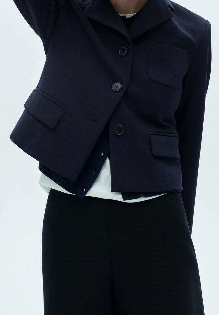 Personne portant un blazer court bleu marine avec boutons et poches à rabat sur un top blanc et un pantalon noir, raccourci au niveau du cou et de la taille.