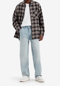 Camisa de cuadros en negro, beige y gris; camiseta blanca debajo; vaqueros sueltos de color azul claro; zapatillas blancas con detalles claros.