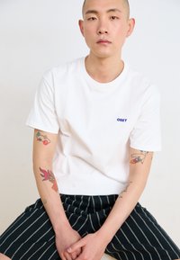 White cotton t-shirt with "OBEY" logo in blue, paired with black pinstriped shorts. Tattoos visible on arms, seated against a plain backdrop.