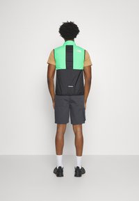 Trackjacke mit grünen und schwarzen Paneelen, beige Hemd darunter, graue Shorts, schwarze Schuhe und weiße Socken, von hinten gesehen.