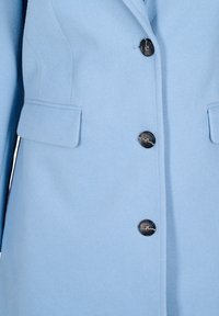 Svetlo modri plašč iz volne z mešanico, ki se ponaša s skrbno oblikovanjem, reverjem, petimi črnimi gumbi in dvema sprednjima žepoma s poklopcem.