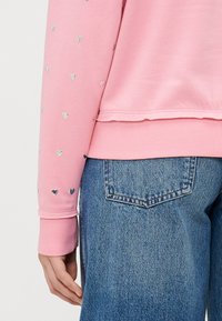 Felpa rosa con decorazioni a cuore argentate sulle maniche, con polsini e fondo a coste, abbinata a jeans di denim blu.