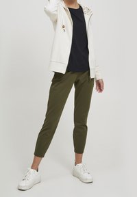 Sudadera blanca con cremallera y acentos florales en la capucha, combinada con pantalones ajustados verde oliva y zapatillas blancas.