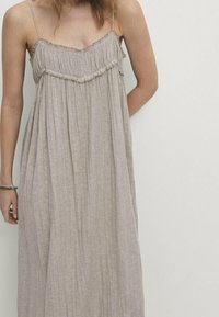 Femme portant une robe maxi plissée beige ample avec des fines bretelles, se tenant devant un fond clair.