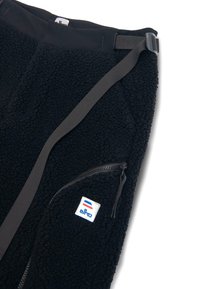 Czarne spodnie polarowe z teksturowaną powierzchnią, regulowany czarny pasek w talii, boczna kieszeń na zamek oraz naszywka z logo z akcentami w kolorze czerwonym, białym i niebieskim.
