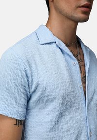 Camisa de manga corta azul claro con una tela texturada, cierre de botones en la parte delantera y un pequeño collar. Tatuajes visibles en el brazo.
