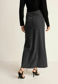 Mujer vistiendo una larga falda de mezclilla negra con dobladillo sin terminar y sandalias de tacón alto negras con tiras, vista desde la espalda contra un fondo liso.