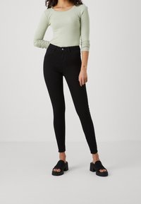 Woman wearing light green ribbed long-sleeve top, high-waisted black skinny jeans, and black heeled sandals standing on a plain background.