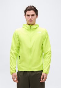 Νεαρός άνδρας που φοράει φωτεινό πράσινο μπουφάν με κουκούλα και φερμουάρ, και σκουρόχρωμο παντελόνι, στέκεται με το πρόσωπο προς τα εμπρός μπροστά σε απλό φόντο.