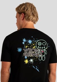 Zwarte katoenen T-shirt met een kleurrijk grafisch ontwerp op de achterkant, inclusief spetters en de tekst "WRONG FRIENDS" met een karakterillustratie.