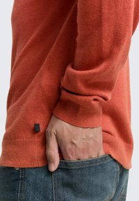 Hand in einer blauen Jeans-Tasche, mit einem orangefarbenen Pulloverärmel, der das Handgelenk bedeckt, und einem kleinen schwarzen Etikett in der Nähe des Saums.