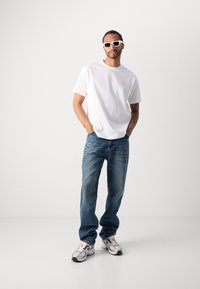 Witte katoenen t-shirt met een klein oranje logo, gecombineerd met blauwe denim jeans. De look wordt compleet gemaakt met witte zonnebril en zilveren sneakers.