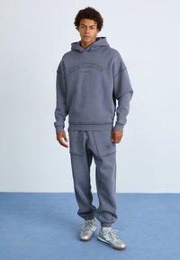 Szara bluza z kapturem i oversize'owym krojem, z detalami w postaci szwów oraz logo. Połączona z pasującymi szarymi spodniami dresowymi.