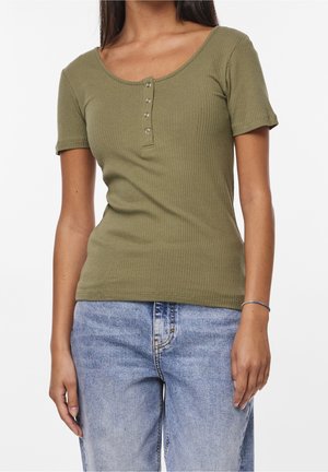 Junge Frau trägt ein olivgrünes, geripptes Henley-Shirt mit kurzen Ärmeln und hellblaue, hoch taillierte Jeans, steht mit entspannten Armen an den Seiten.