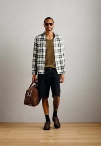 Man walking indoors carrying brown leather duffel bag, wearing sunglasses, checkered shirt, olive tank top, black shorts, and brown shoes.
