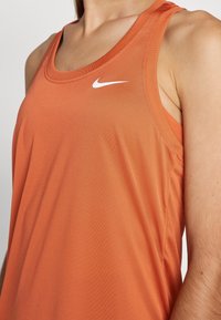Close-up van een persoon die een oranje mouwloos Nike-shirt draagt met een wit logo op de borst.